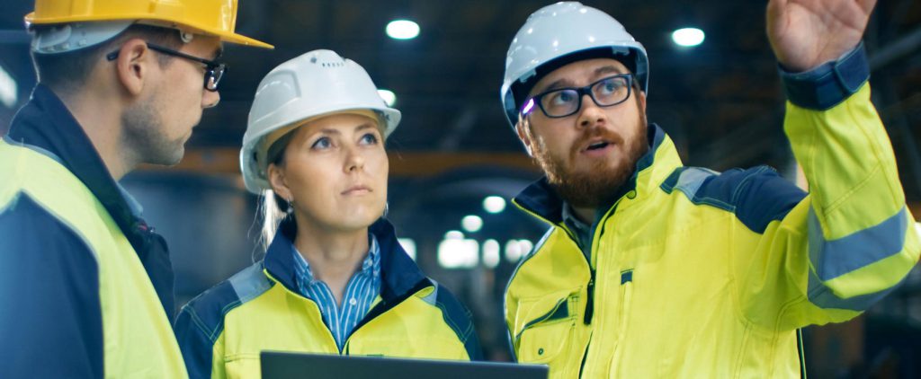 Two men and a woman, wearing white helmets and neon yellow and gray uniforms, are standing, and one of the men is talking.