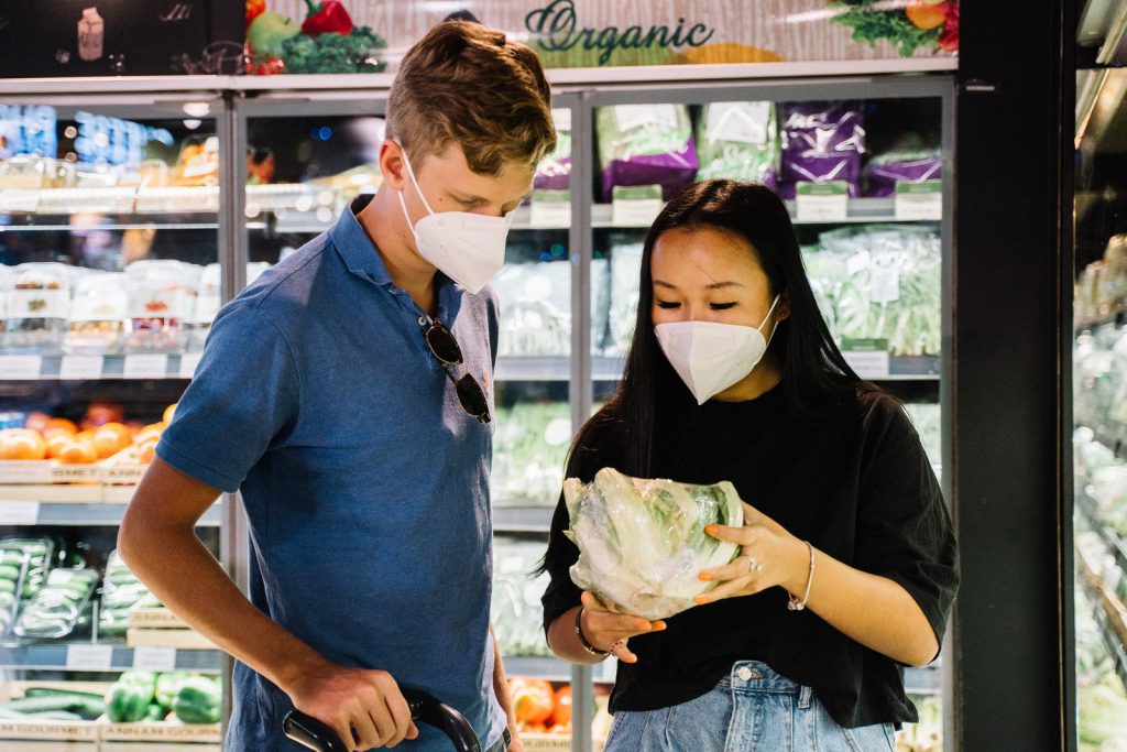 A woman and a man wearing masks are standing in front of cold vegetable cabinets in a grocery store, looking at a vegetable.