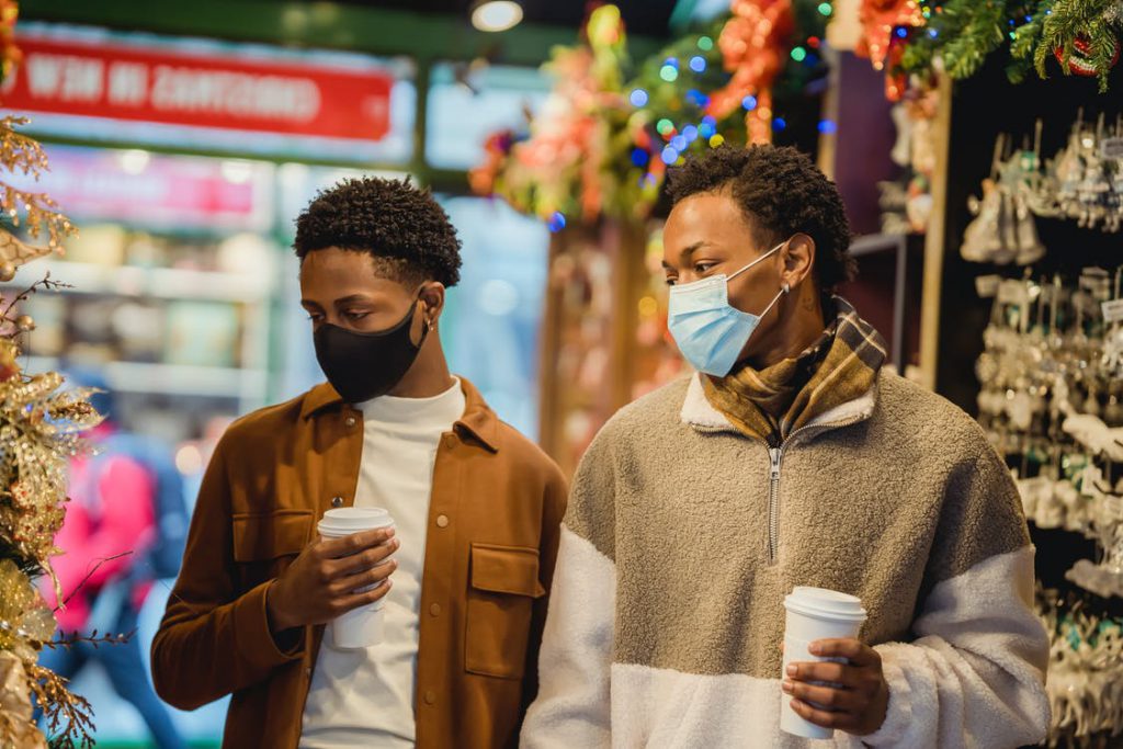 Two men wearing blue and black masks are standing with take away coffee in their hands, looking at the Christmas decorations.