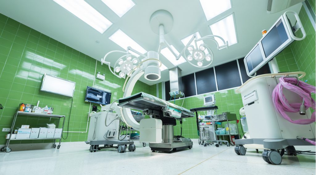 Surgical supplies and equipment, operating table, screens and lights in an empty operating room with green ceramic walls.