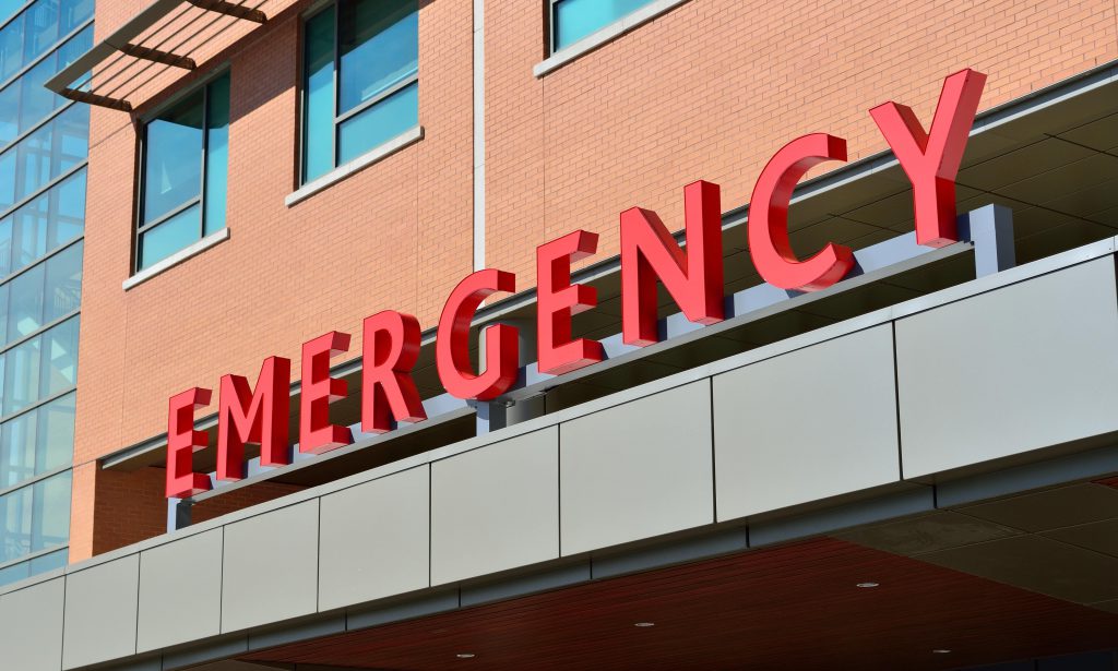 Yurtdışında bir hastane binasının acil servis kısmının dışarıdan görüntüsü, kırmızı büyük harflerle "EMERGENCY" yazıyor. 