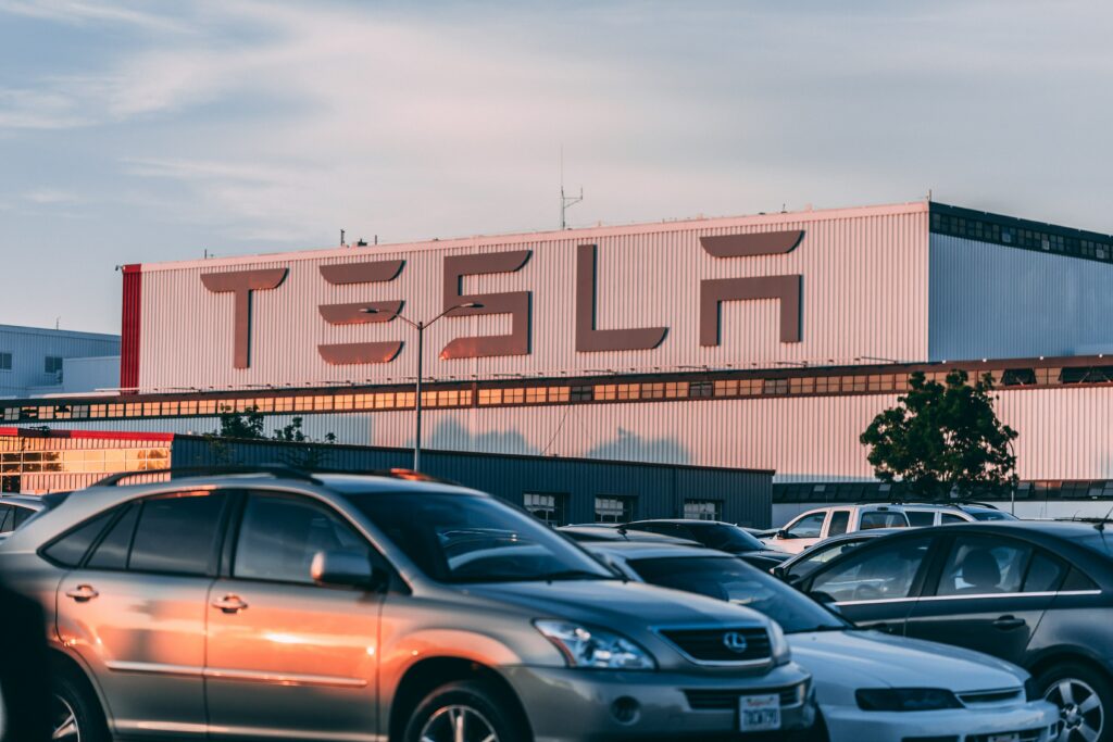 Tesla fabrikasının önüne park edilmiş arabalar ve büyük harflerle TESLA logosu.